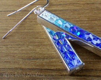 May include: Silver bar earrings filled with blue and clear crystals. The crystals transition from light blue to deep blue and purple. The earrings have a hook closure. The image is on a wooden surface.