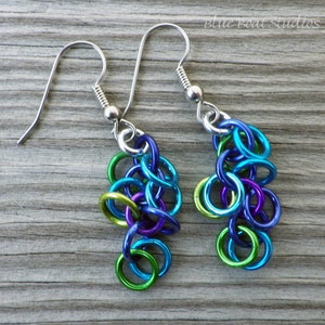 May include: A pair of dangle earrings with a cluster of small, colorful metal rings. The rings are in shades of blue, green, and purple. The earrings are made with silver-toned metal.