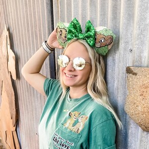 May include: A green t-shirt with a pocket featuring Simba and Nala from The Lion King. The text on the pocket reads "Can you feel the love tonight". The person wearing the shirt is also wearing green glitter mouse ears with a bow.
