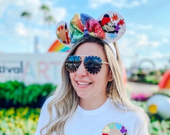 Op de afbeelding: Een wit T-shirt met een kleurrijke verfpalette-zak en de tekst "Imagination has no limit". De persoon draagt een regenboog glitter hoofdband met Mickey Mouse oren.