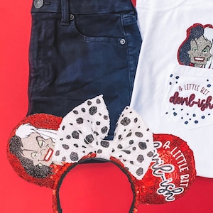 May include: A white t-shirt with a red and black embroidered image of Cruella de Vil and the text "A little bit devil-ish". The shirt is paired with dark blue jeans and a red and white polka dot headband with a Cruella de Vil image.
