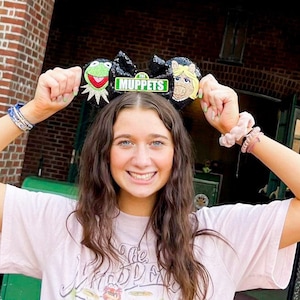 Puede incluir: Una persona joven con una camiseta rosa con un gráfico de la banda de los Muppets y una diadema negra con Kermit la Rana, Miss Piggy y un cartel de calle que dice "Muppets". La persona está de pie frente a un edificio de ladrillo con un cartel que dice "Now Playing Muppet Vision".