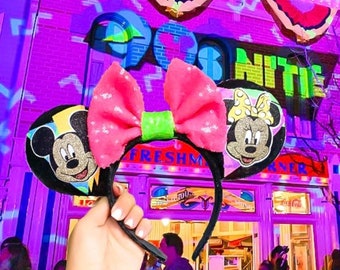 May include: A pair of pink and black Minnie Mouse ears with a green bow. The ears are in front of a building with the words "Goofy's Nittie Knittie" on the side.