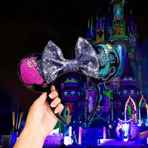 Puede incluir: Un par de orejas de Minnie Mouse negras con un lazo de purpurina plateado. Las orejas presentan un personaje rosa y morado en una oreja y un personaje azul y verde en la otra oreja. Las orejas están frente a un fondo oscuro con un castillo iluminado en la distancia.
