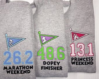 May include: Three grey sweatshirts with different Disney-themed designs. The first sweatshirt has a blue flag with Mickey Mouse and the text "26.2 Marathon Weekend". The second sweatshirt has a green flag with Dopey and the text "48.6 Dopey Finisher". The third sweatshirt has a pink flag with a castle and the text "13.1 Princess Weekend".
