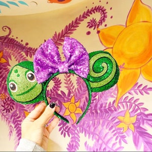 Peut inclure: Oreilles de Minnie Mouse en sequins verts et violets avec un motif de grenouille sur une oreille et un motif de spirale sur l'autre.