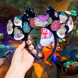 May include: Black Minnie Mouse ears with a sparkly black background and colourful gems. The ears are decorated with images of the seven dwarfs from the Disney movie Snow White and the Seven Dwarfs.