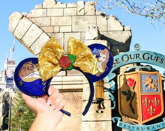 May include: A pair of blue and gold Minnie Mouse ears with a gold bow and a red rose. The ears feature Belle and the Beast from Disney's Beauty and the Beast. The ears are in front of a sign that says "Be Our Guest Restaurant".