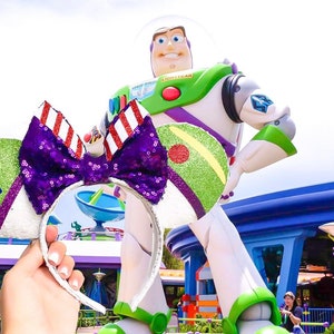 May include: A pair of green and purple Minnie Mouse ears with a red and white striped bow. The ears are decorated with green, red, and white details. The ears are in front of a large statue of Buzz Lightyear from the Toy Story franchise.