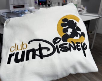 May include: White sweatshirt with the embroidered text "club runDisney" in black and gold. The design includes a black silhouette of Mickey Mouse inside a gold letter "C" and the word "Disney".