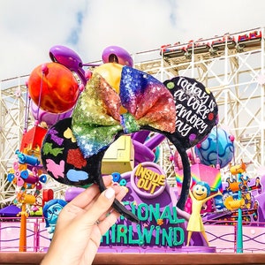 May include: A pair of black mouse ears with a rainbow sequin bow and a black felt circle with the text "Today is a core memory day" in white. The ears are in front of a colourful amusement park background.