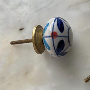 Puede incluir: Un pomo de cer&aacute;mica blanca con un dise&ntilde;o floral azul y rojo. El pomo est&aacute; unido a un tornillo de metal dorado.