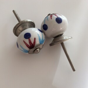 Puede incluir: Dos pomos de armario de cer&aacute;mica blanca con dise&ntilde;os pintados en azul y rojo. Los pomos tienen una base de metal plateado y un tornillo.