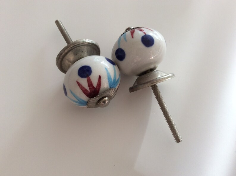 Puede incluir: Dos pomos de armario de cer&aacute;mica blanca con dise&ntilde;os pintados en azul y rojo. Los pomos tienen una base de metal plateado y un tornillo.