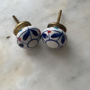 Puede incluir: Dos pomos de cer&aacute;mica blanca con dise&ntilde;os florales azules y rojos. Los pomos tienen una base de metal dorado.