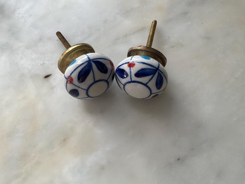 Puede incluir: Dos pomos de cer&aacute;mica blanca con dise&ntilde;os florales azules y rojos. Los pomos tienen una base de metal dorado.
