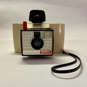 May include: Vintage Polaroid Swinger Model 20 Land Camera. The camera has a black and silver face with a large lens and the words "Polaroid Land Camera Swinger Model 20" printed on the front. It has a cream-colored body, a black viewfinder, and a red button.