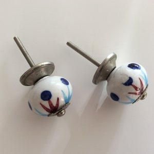 Puede incluir: Dos pomos de armario de cer&aacute;mica blanca con un dise&ntilde;o floral azul y rojo. Los pomos tienen una base de metal plateado y un tornillo.
