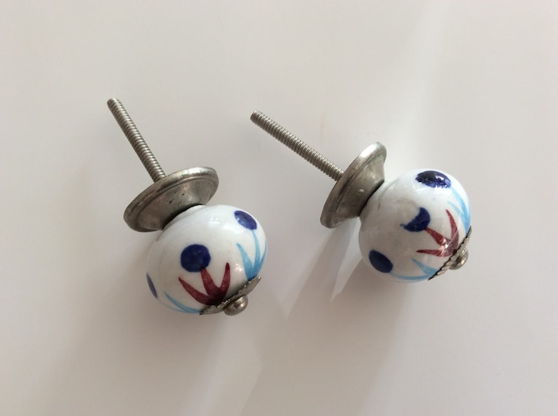 Puede incluir: Dos pomos de armario de cer&aacute;mica blanca con un dise&ntilde;o floral azul y rojo. Los pomos tienen una base de metal plateado y un tornillo.