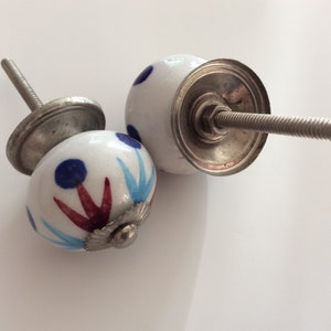 Puede incluir: Dos pomos de armario de cer&aacute;mica blanca con dise&ntilde;os pintados en azul y rojo. Los pomos tienen una base de metal plateado y un tornillo.
