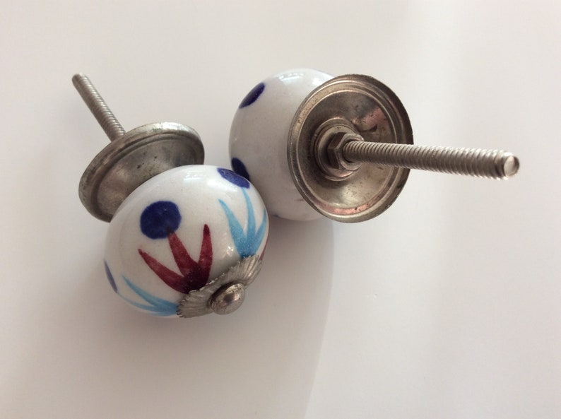 Puede incluir: Dos pomos de armario de cer&aacute;mica blanca con dise&ntilde;os pintados en azul y rojo. Los pomos tienen una base de metal plateado y un tornillo.