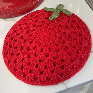 Puede incluir: Un salvamanteles de ganchillo rojo hecho a mano con forma de fresa, con un tallo verde. El salvamanteles redondo está sobre una encimera blanca. Un electrodoméstico de cocina rojo está en el fondo.