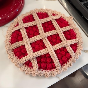 May include: A handmade, round crochet trivet resembling a cherry pie. The trivet features a red filling, a tan lattice crust, and a scalloped edge. This decorative item is designed to protect surfaces from hot dishes.