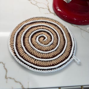 May include: A round, crocheted trivet with a spiral design. The trivet features concentric circles of brown, tan, and white yarn. It has a small loop for hanging. This handmade item is designed for kitchen use.