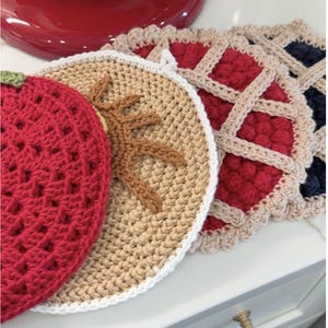 Könnte beinhalten: Eine Sammlung handgefertigter gehäkelter Topflappen. Sie zeigen Designs wie eine rote Erdbeere mit grünem Stiel, ein beigefarbenes Exemplar mit braunem Blattmuster und Tortenstücke mit roter und marineblauer Füllung und Gitterkrusten.