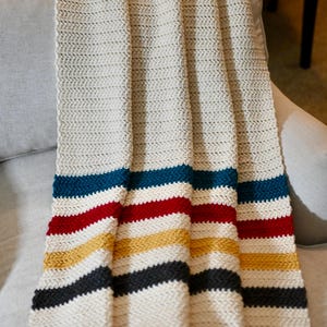 May include: Cream-colored knitted blanket with horizontal stripes in blue, red, yellow, and black. The blanket is draped over a light gray armchair, showcasing its textured weave and vibrant color scheme. A cozy home decor item.