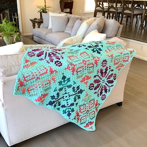 May include: A teal crochet throw blanket with a colorful geometric pattern. The blanket is draped over a light gray couch.