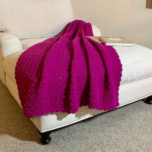 May include: A soft, pink crocheted throw blanket draped over a white armchair. The blanket has a textured, woven pattern.