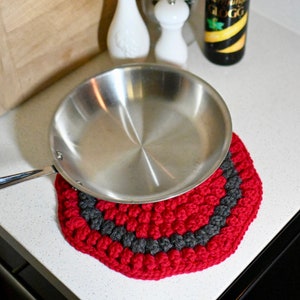 以下が含まれることがあります： 赤とグレーのクロシェの鍋敷きに、ステンレス製のフライパンが置かれています