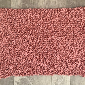 Op de afbeelding: Een rechthoekig, handgemaakt tapijt in een stoffige roze kleur. Het tapijt heeft een getextureerd, gelust oppervlak, waardoor het een zachte uitstraling heeft. Het tapijt is waarschijnlijk gemaakt van garen of een vergelijkbaar materiaal en is geschikt voor een badkamer of slaapkamer.