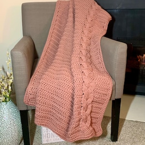 Op de afbeelding: Een zachte, roze gehaakte deken met een kabelbreipatroon, gedrapeerd over een stoel. De deken is in een warme, neutrale kleur en perfect om elke kamer een gezellige sfeer te geven.