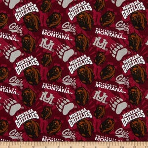 May include: A maroon fabric featuring a repeating pattern of brown grizzly bear heads, white bear paw prints, and the text "Montana Grizzlies" and "University of Montana" in white, with a ruler visible at the bottom edge showing 11 inches.