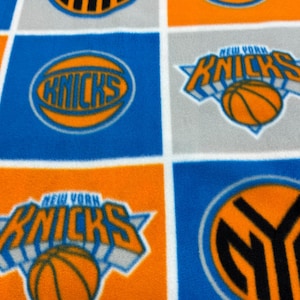 May include: A fabric with a repeating pattern of New York Knicks logos. The logos include the team name, a basketball, and the letters "NYK" in orange, blue, and white. The fabric is divided into squares.
