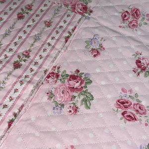 May include: A quilted fabric featuring a pink background with a floral pattern of red roses and small blue flowers. The design includes a striped border with pink and white stripes and floral accents. The fabric has a diamond-stitched pattern.