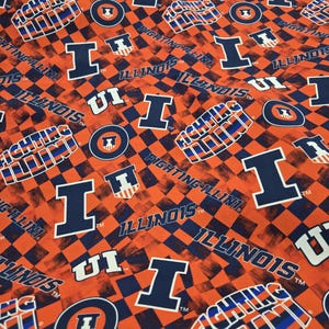 May include: A fabric pattern featuring a repeating orange and dark blue checkerboard background with the text Illinois, Fighting Illini, and the letter I logo in white and dark blue, displaying official university sports team branding throughout.