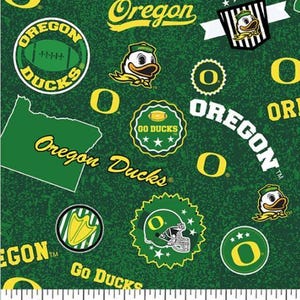 May include: Green fabric featuring a repeating pattern of Oregon Ducks logos, text, and football imagery. A white ruler at the bottom shows a scale of 7 inches, indicating the print size of the various team graphics and symbols on the material.