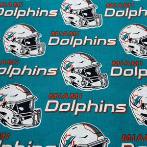 May include: A teal fabric featuring a repeating pattern of white Miami Dolphins football helmets with orange and teal logos, paired with the text Miami Dolphins in white letters with orange outlines, arranged in a staggered grid layout.