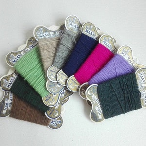 Może przedstawiać: Zestaw kolorowych szpul z włóczką w różnych odcieniach zieleni, brązu, niebieskiego, różowego, fioletowego i szarego. Każda szpula jest owinięta w białą kartkę z nadrukiem "MAAS" i "SCHEEPSWOL".