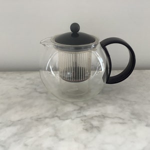 Puede incluir: Una tetera de vidrio transparente con tapa y asa negras. La tetera tiene un filtro integrado para té de hojas sueltas.