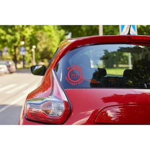 May include: Red car with a circular sticker on the rear window. The sticker features a red gear shift graphic with the words "MILLENNIAL ANTI-THEFT DEVICE" in a circular pattern around the graphic.