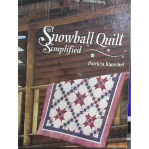 May include: A book cover titled "Snowball Quilt Simplified" by Patricia Knoechel. The cover features a quilt with white circles and red starbursts on a blue background. The quilt is displayed on a wooden porch railing.