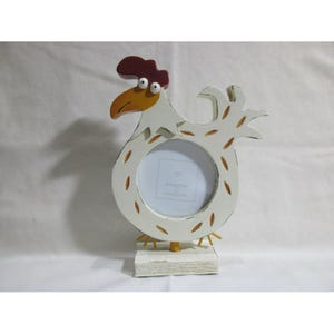 Può includere: Una cornice a forma di gallo bianca con cresta rossa e becco giallo. La cornice ha un'apertura circolare per una foto ed è montata su una base di legno bianca. La cornice ha accenti marroni e il testo "SONOMA" è visibile.