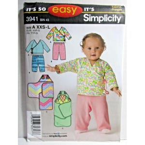 Pode incluir: Um envelope de molde de costura Simplicity, número 3941, com ilustrações de roupas de bebê, incluindo um top transpassado, calças e uma manta de embrulho. A embalagem mostra uma criança vestindo um top floral e calças cor-de-rosa.