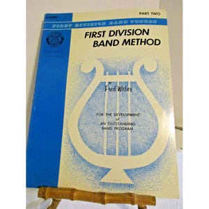 Puede incluir: Un libro de música vintage titulado "First Division Band Method" de Fred Weber. La portada presenta una combinación de colores azul y crema con un gráfico de lira. El texto incluye "Part Two" y "For the Development of an Outstanding Band Program."