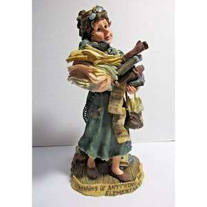 May include: A figurine of a woman wearing glasses and a green dress, holding a stack of papers and books. The base of the figurine has the text "Teaching is ANYTHING but ELEMENTARY!"