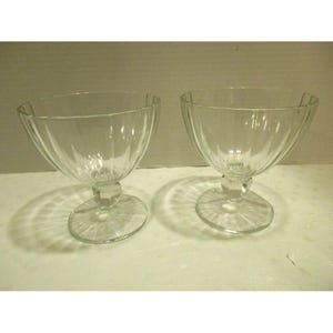 May include: Two clear glass dessert bowls with a ribbed design. Each bowl has a rounded base and a stem that connects to a circular foot. The bowls are empty and sit on a white surface.